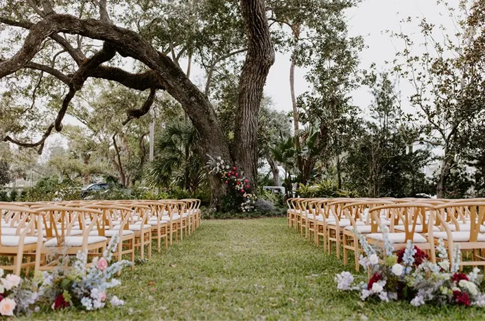 Outdoor ceremony setup with wooden chairs arranged under a large tree, accented by lush greenery and soft floral arrangements