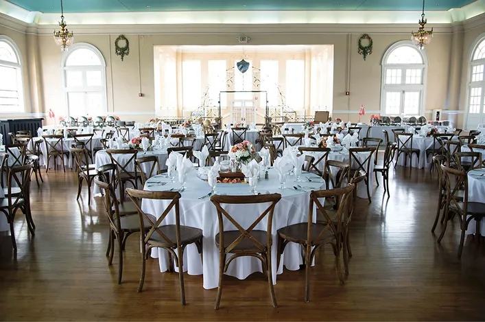 Elegant reception space with round tables, wooden chairs, white linens, floral centerpieces, and large arched windows
